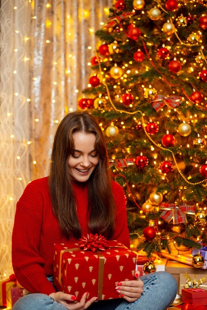 Happy smiling girl sitting near decorated Christmas tree with present box in her hands Magical happy time of winter holidays and warm atmosphere