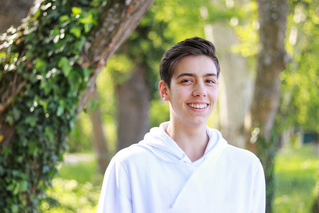 Happy smiling handsome teenager boy in white hoody in park outdoors at bright sunny day