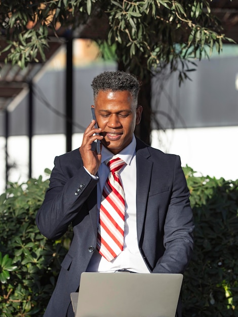 A happy smiling Hispanic businessman talking on a smartphone and working from his laptop remotely