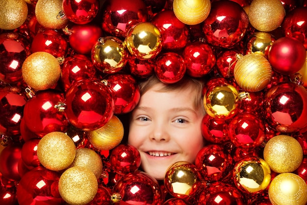 Happy Smiling Kid with Christmas Balls Toys. Child over Red and Golden Bauble Background. Shining Bright Xmas Tree Holiday Decoration