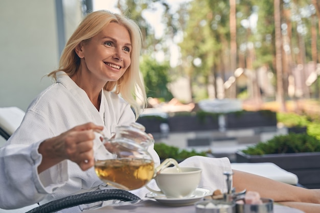 Happy smiling lady holding teapot in hand while poring tea in white cup at the desk