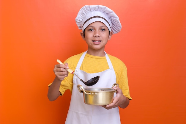 Happy smiling little boy in apron with saucepan and soup spoon  over orange background