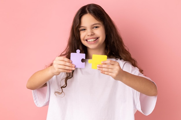 Happy smiling little girl holding yellow and purple puzzle pieces solving task looking at camera