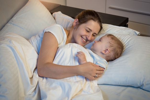 Happy smiling mother embracing her baby in bed at night