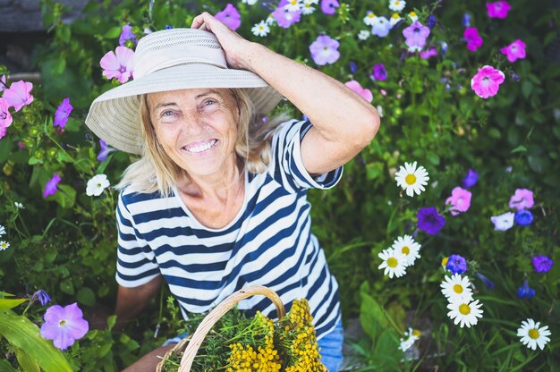 Happy smiling senior woman posing in summer garden with flowers in straw hat.