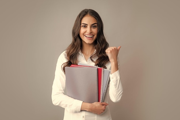 Happy smiling student or teacher Teen girl with notebooks standing over gray background with copy space Educational concept for school Portrait of young student Winner sign