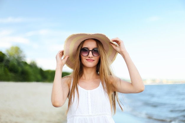 Happy smiling woman in free happiness bliss on ocean beach standing and posing with hat and sunglasses. Portrait of a female model in white summer dress enjoying nature during travel holidays vacation