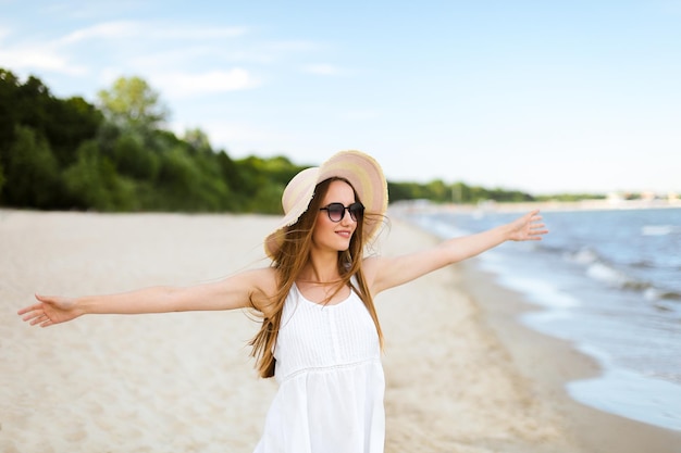 Happy smiling woman in free happiness bliss on ocean beach standing with a hat, sunglasses, and open hands. Portrait of a multicultural female model in white summer dress enjoying nature during travel