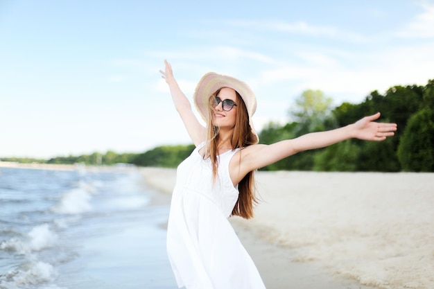 Happy smiling woman in free happiness bliss on ocean beach standing with a hat, sunglasses, and rasing hands. Portrait of a multicultural female model in white summer dress enjoying nature during trav