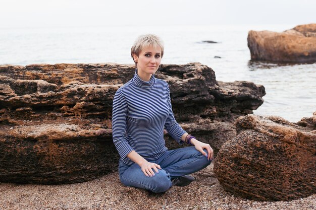 Happy smiling woman sitting on the rock near sea in autumn or summer time