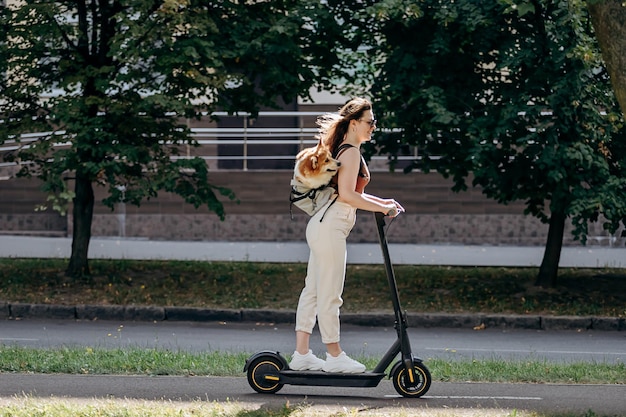 Photo happy smiling woman traveler is riding her electro scooter in city parkland with dog welsh corgi pembroke in a special backpack