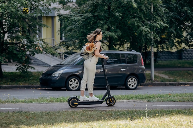 Happy smiling woman traveler is riding her electro scooter in city parkland with dog Welsh Corgi Pembroke in a special backpack