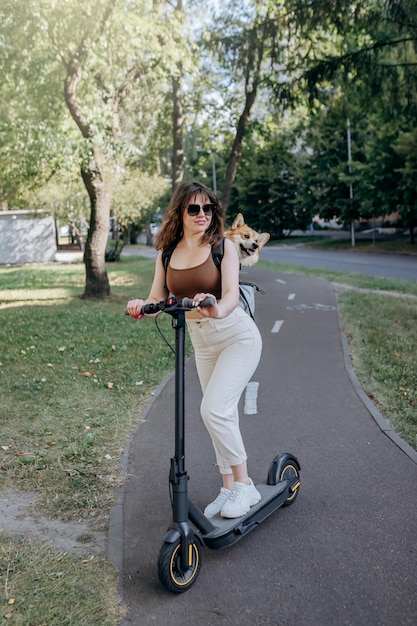 Happy smiling woman traveler is riding her electro scooter in city parkland with dog Welsh Corgi Pembroke in a special backpack