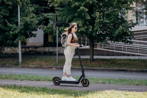 Happy smiling woman traveler is riding her electro scooter in city parkland with dog Welsh Corgi Pembroke in a special backpack