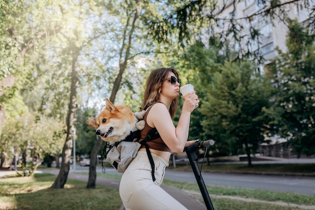 Happy smiling woman traveler is standing with electro scooter and drinking coffee in city parkland with dog Welsh Corgi Pembroke in a special backpack