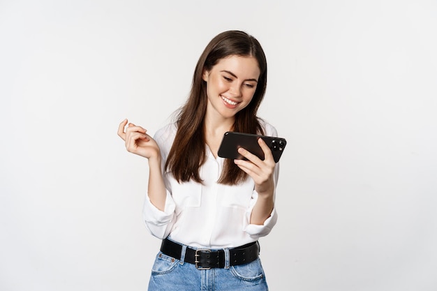 Happy smiling woman watching on mobile phone, looking at smartphone and laughing, standing over white background.