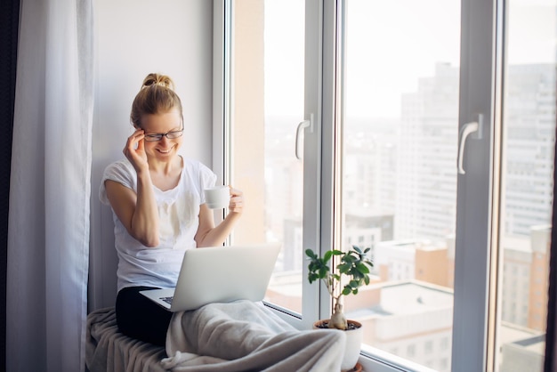 Happy smiling woman wearing glasses sitting on windowsill at city apartment, resting, looking at webcam on laptop, talking on video chat with her friends or family.