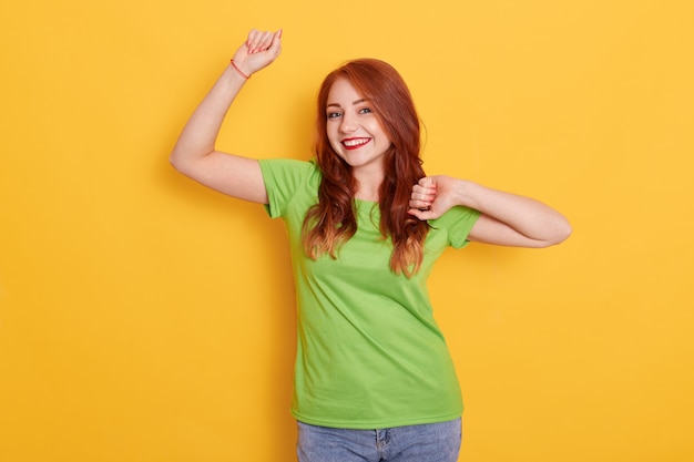 Happy smiling woman wearing green casual t shirt raising hands up, celebrating victory and dancing, posing isolated over yellow background, looks happy and joyful.