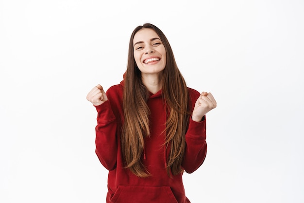 Happy smiling woman winning and celebrating, look delighted with fist pumps, say yes, achieve goal, feeling sweet taste of success, become champion, standing over white wall