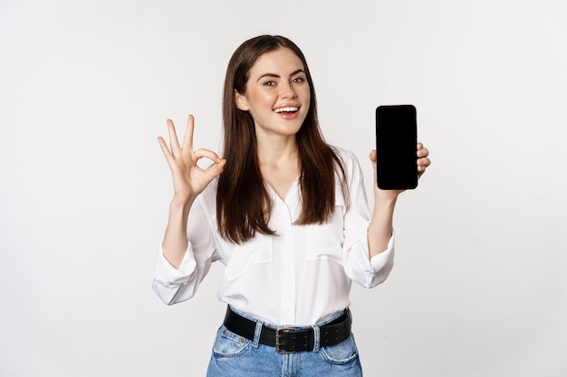 Happy smiling woman with satisfied face, recommending excellent app, showing mobile phone screen, okay sign, standing over white background.