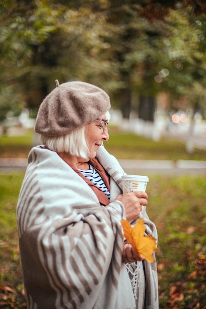 Happy smiling woman with yellow maple leaves coffee cup in french beret walking in autumn park