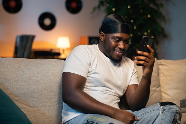 Happy smiling young africanamerican man with bandana on head and white tshirt sits on sofa at home