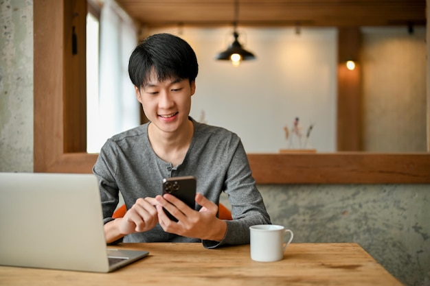 Happy and smiling young Asian man using his smartphone at a table in the coffee shop