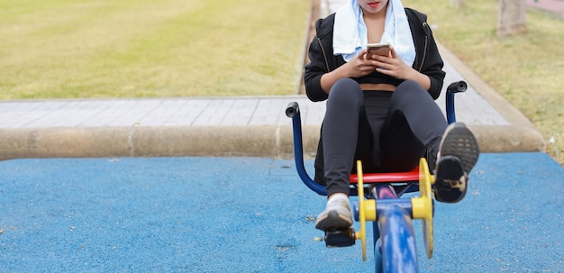 Happy smiling young beautiful asian woman in sportswear relaxing on public exercise machine. Cute and sporty girl working out on bike and using mobile smartphone outdoor