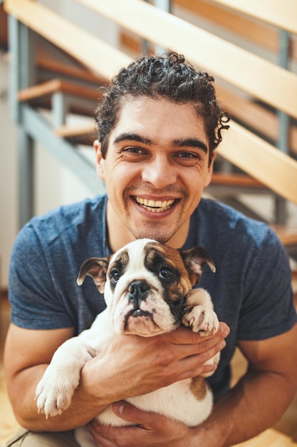 A happy smiling young man is sitting on a floor in the living room of his new apartment and holding a little puppy.