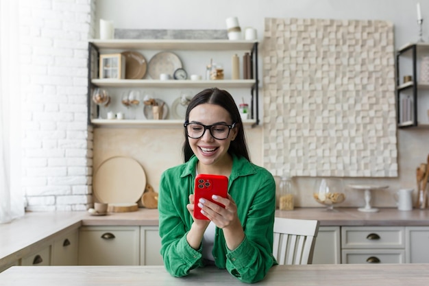 Happy and smiling young teenage girl sittiing at home in the kitchen and using the phone receives