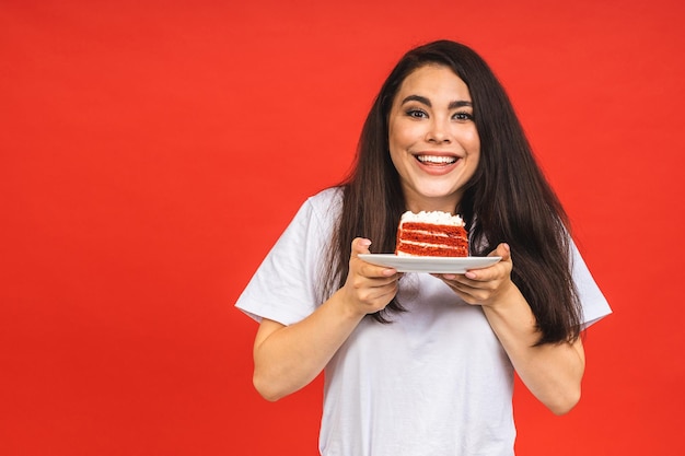 Happy smiling young woman eating the cake isolated over red background Brunette lady holding a birthday cake