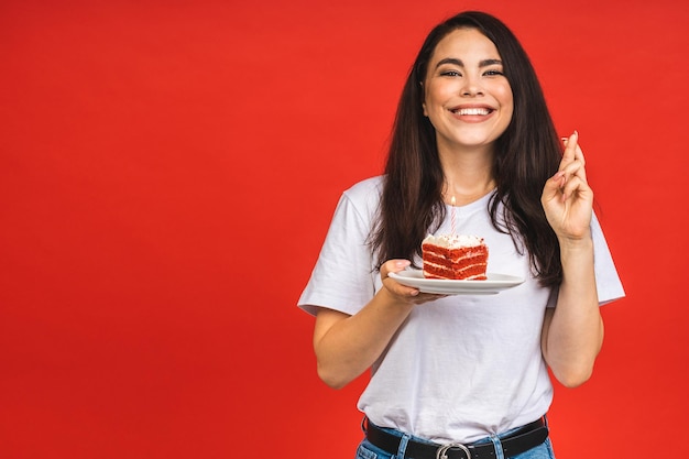 Happy smiling young woman eating the cake isolated over red background Brunette lady holding a birthday cake