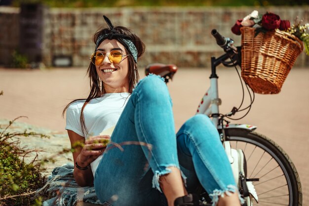 Happy smiling young woman is enjoying in a summer sunny day reposing on little wall in a city, beside the bike with flower basket.