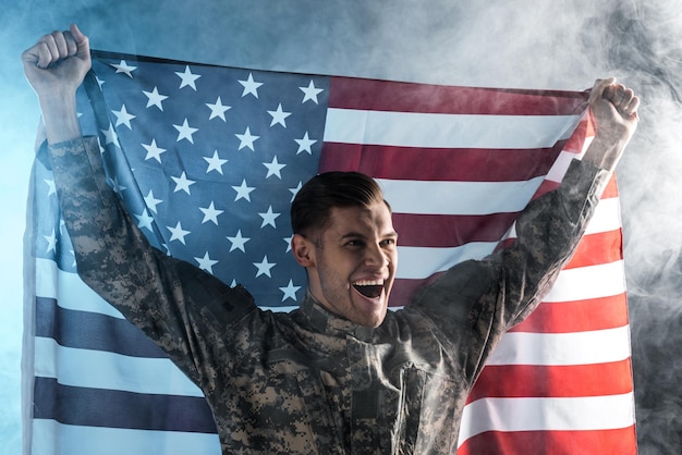 Photo happy soldier gesturing while holding american flag on black with smoke