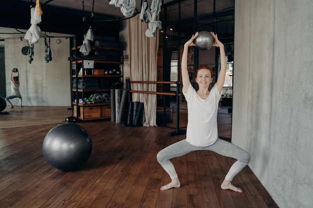 Happy sportive female in active wear holding mini fitball above head in half sitting position