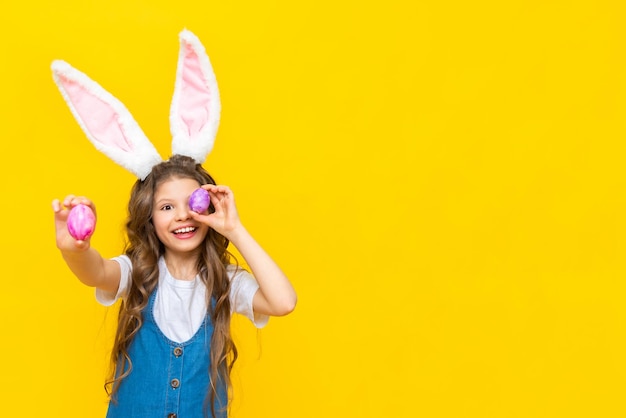 Happy spring Easter The little girl closes her eyes with eggs and smiles broadly A charming child with rabbit ears in a blue sundress on a yellow isolated background Copy space