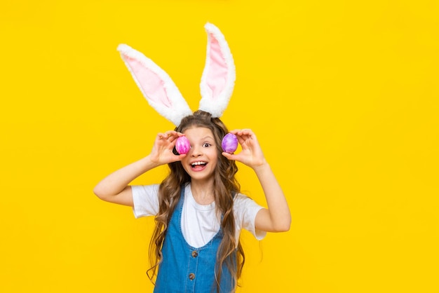 Happy spring Easter The little girl closes her eyes with eggs and smiles broadly A charming child with rabbit ears in a blue sundress on a yellow isolated background