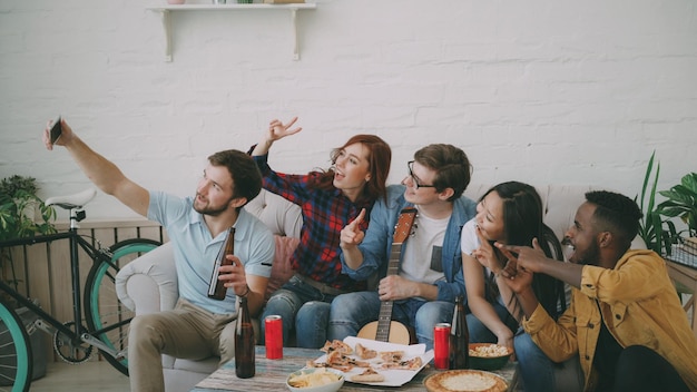 Happy student friends taking selfie on smartphone and posing while have party with beer and pizza in shared accommodation indoors