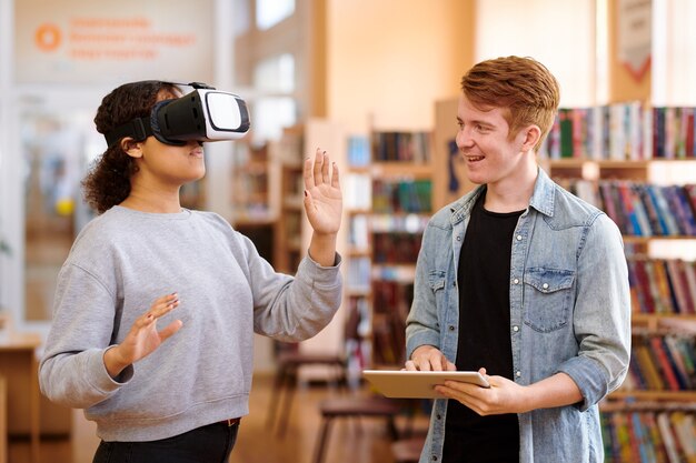 Happy student using tablet while interacting with his classmate with vr headset in library