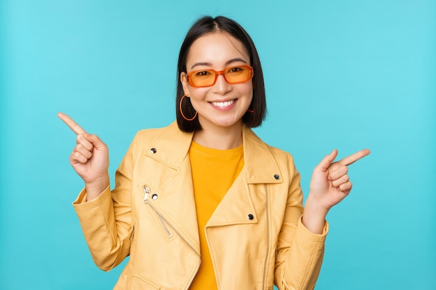 Happy stylish chinese girl in sunglasses points fingers sideways invites to check out see store discount shows left and right stands over blue background