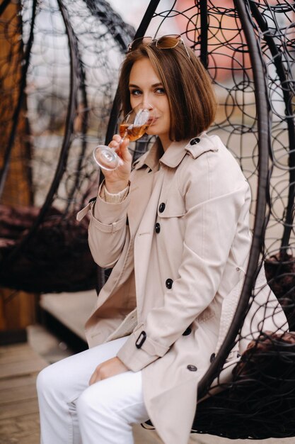 A happy stylish girl In a gray coat is sitting outside in an armchair and drinking a drink