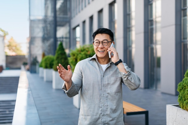 Happy successful asian businessman talking on smartphone standing outdoors against modern office center free space