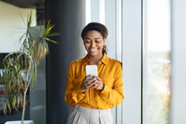 Happy successful black businesswoman typing on smartphone standing near window in modern office interior