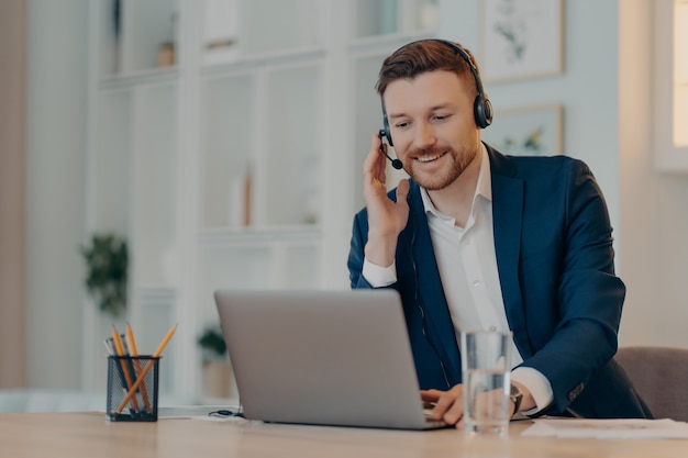 Happy successful businessman using laptop and typing on keyboard while sitting at the table and doing video call with headset, taking part in web conference. Online business concept