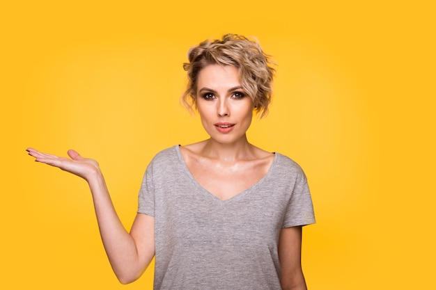 Happy surprised blonde young female smiling broadly at camera pointing fingers away showing something interesting and exciting on yellow studio background with copy space for your text