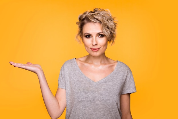 Happy surprised blonde young female smiling broadly at camera pointing fingers away showing something interesting and exciting on yellow studio background with copy space for your text