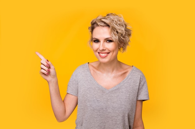 Happy surprised blonde young female smiling broadly at camera pointing fingers away showing something interesting and exciting on yellow studio background with copy space for your text