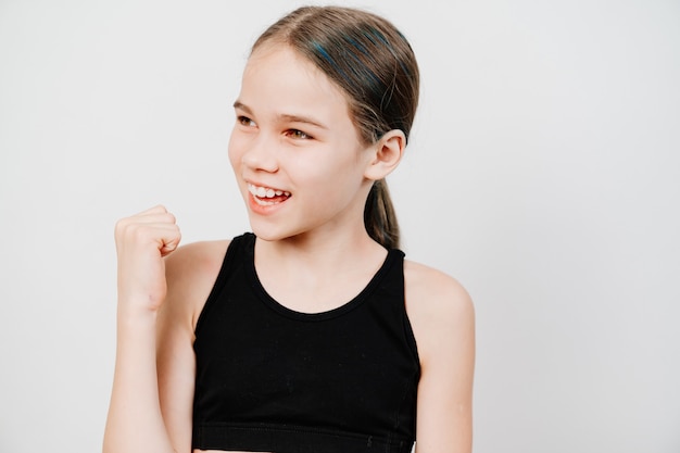 Happy teen girl in a black T-shirt stands on a white surface and smiles showing teeth.