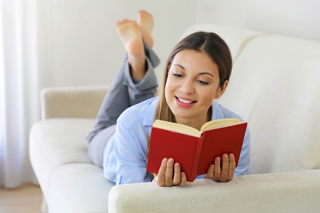 Happy teen girl lying on sofa in the living room while reading her favorite author of books