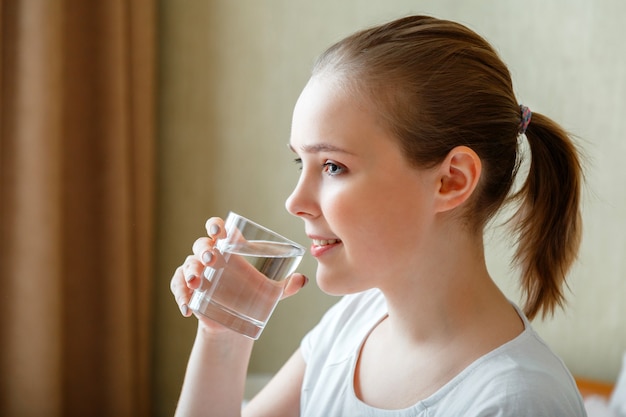 Happy teen girl maintains water balance for body health by drinking transparent cup of clean water. Smile young woman drinks glass of pure water in morning after waking up.